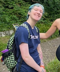 man with dirty blond short hair, wearing blue sunglasses and a grey top and white skirt, has some facial piercings